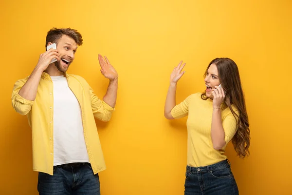 Casal alegre acenando as mãos um para o outro enquanto fala em smartphones no fundo amarelo — Fotografia de Stock