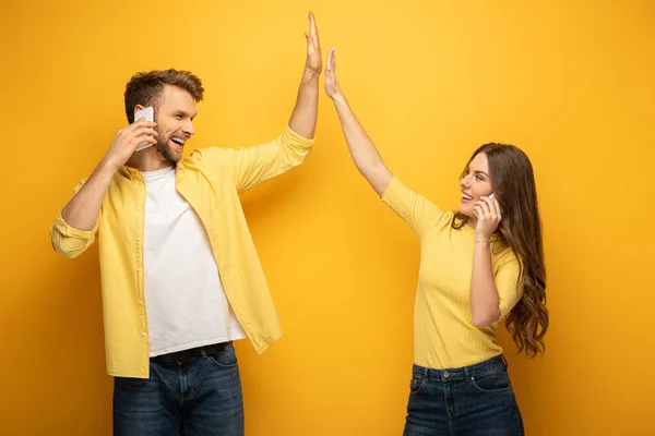 Alegre pareja alta cinco y sonriendo el uno al otro mientras se habla en los teléfonos inteligentes en el fondo amarillo - foto de stock