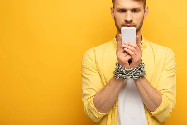 Bel homme avec chaîne métallique autour des mains tenant smartphone sur fond jaune — Photo de stock