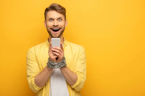 Homme joyeux avec chaîne métallique autour des mains tenant smartphone sur fond jaune — Photo de stock