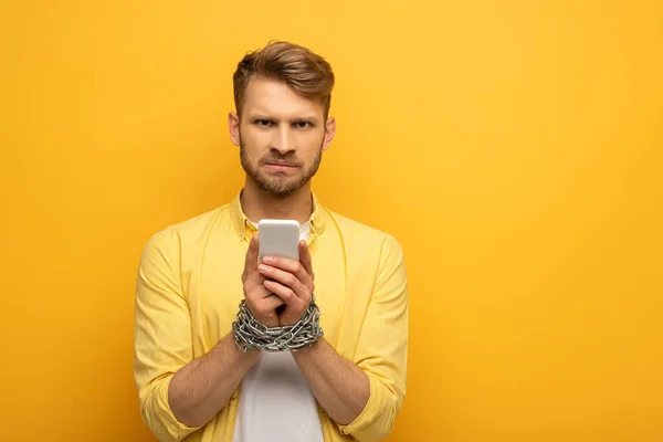 Sad man with tied hands with metal chain holding smartphone on yellow background — Stock Photo