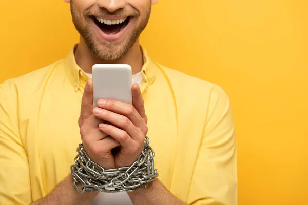 Vista recortada del hombre feliz con cadena alrededor de las manos sosteniendo el teléfono inteligente sobre fondo amarillo - foto de stock