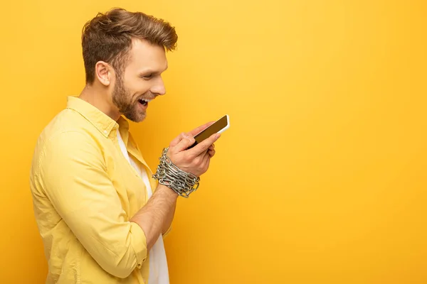 Вид сбоку улыбающегося человека с цепью вокруг рук, держащего смартфон на желтом фоне — стоковое фото