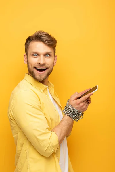 Side view of cheerful man with metal chain around hands holding smartphone and looking at camera on yellow background — Stock Photo