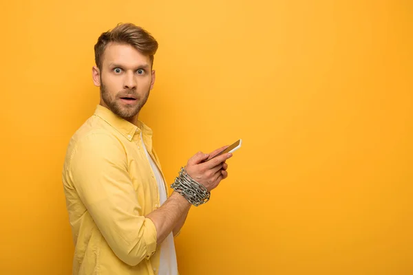 Vista lateral do homem confuso com corrente de metal em torno de mãos segurando smartphone e olhando para a câmera no fundo amarelo — Fotografia de Stock