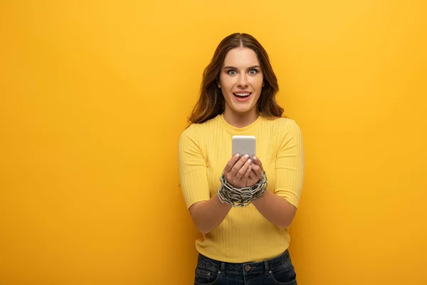 Schöne lächelnde Frau mit gefesselten Händen mit Metallkette, die Smartphone auf gelbem Hintergrund hält — Stockfoto