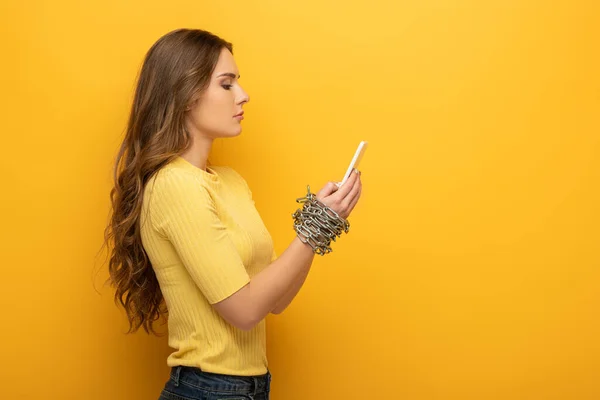 Vista lateral de mujer atractiva sosteniendo teléfono inteligente en manos atadas con cadena de metal sobre fondo amarillo - foto de stock