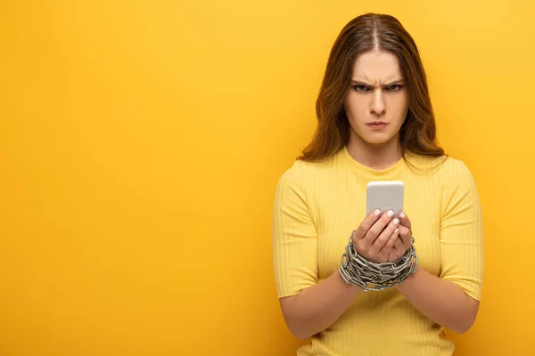 Wütendes Mädchen blickt in die Kamera, während es sein Smartphone in gebundenen Händen mit Kette auf gelbem Hintergrund hält — Stockfoto