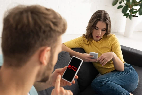 KIEW, UKRAINE - 21. FEBRUAR 2020: Selektiver Fokus des Mannes mittels netflix-App auf dem Smartphone in der Nähe von Freundin, die auf der Couch chattet — Stockfoto