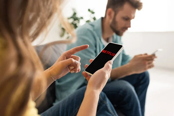 KYIV, UKRAINE - FEBRUARY 21, 2020: Selective focus of woman holding smartphone with netflix app near boyfriend on couch — Stock Photo