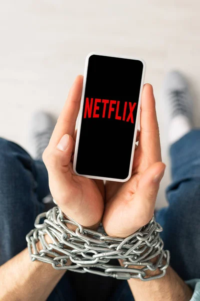 KYIV, UCRANIA - 21 de febrero de 2020: Vista superior del hombre sosteniendo el teléfono inteligente con la aplicación netflix en manos atadas con cadena de metal - foto de stock