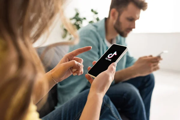 KYIV, UKRAINE - FEBRUARY 21, 2020: Selective focus of woman pointing with finger at smartphone with TikTok app near boyfriend chatting on couch — Stock Photo