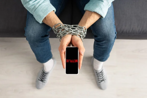 KYIV, UKRAINE - FEBRUARY 21, 2020: Top view of man with metal chain around hands holding smartphone with netflix app on couch — стокове фото