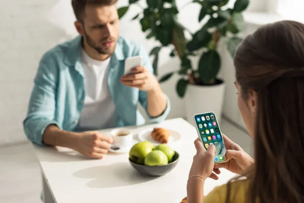 KYIV, UCRANIA - 21 de febrero de 2020: enfoque selectivo de la mujer que sostiene el teléfono inteligente con pantalla de iphone cerca de su novio charlando durante el desayuno - foto de stock