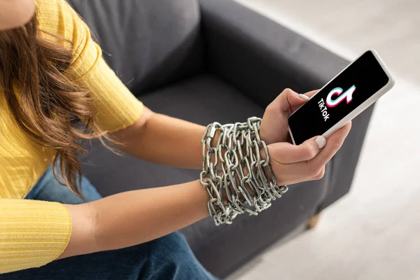 KYIV, UKRAINE - FEBRUARY 21, 2020: Cropped view of young woman holding smartphone with TikTok app in tied hands with metal chain on couch — Stock Photo