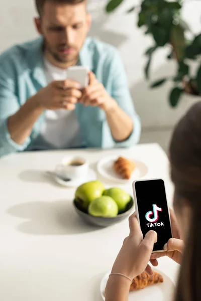 KYIV, UCRANIA - 21 de febrero de 2020: enfoque selectivo de la niña sosteniendo el teléfono inteligente con la aplicación TikTok cerca del novio durante el desayuno - foto de stock