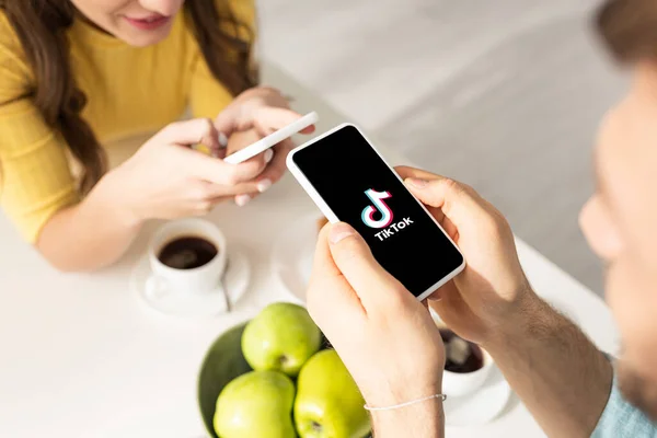 KYIV, UKRAINE - FEBRUARY 21, 2020: Selective focus of man holding smartphone with TikTok app near girl and coffee on table — Stock Photo