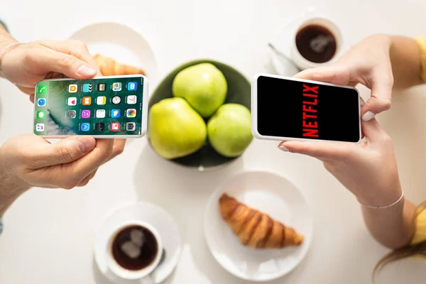 KYIV, UKRAINE - FEBRUARY 21, 2020: Cropped view of couple holding smartphones with iphone screen and netflix app near breakfast on table — Stock Photo