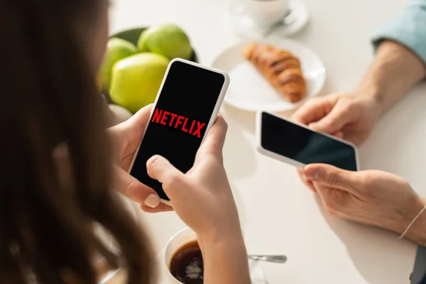 KYIV, UCRANIA - 21 DE FEBRERO DE 2020: Enfoque selectivo de la niña usando el teléfono inteligente con la aplicación de netflix cerca del novio y el desayuno en la mesa - foto de stock