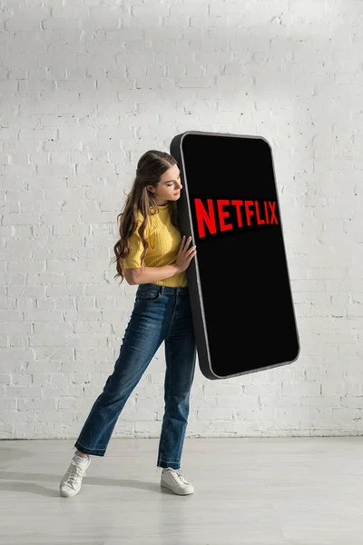 KYIV, UKRAINE - FEBRUARY 21, 2020: Beautiful woman holding huge model of smartphone with netflix app near white brick wall — Stock Photo