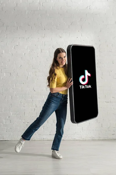 KYIV, UKRAINE - FEBRUARY 21, 2020: Beautiful girl smiling and looking at camera while holding big model of smartphone with TikTok app — Stock Photo