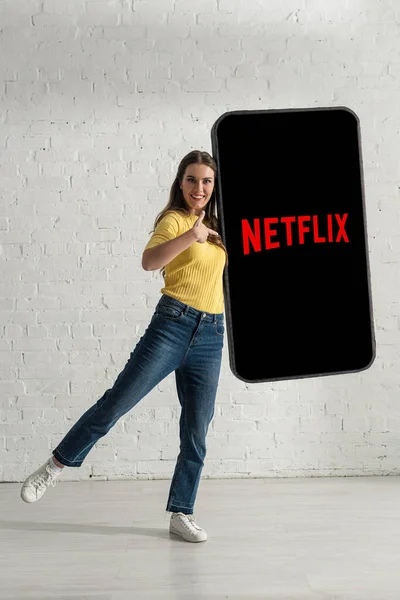 KYIV, UCRANIA - 21 de febrero de 2020: Muchacha sonriente que señala con el dedo al modelo de teléfono inteligente con aplicación netflix cerca de la pared de ladrillo blanco — Stock Photo