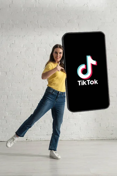 KYIV, UKRAINE - FEBRUARY 21, 2020: Beautiful young woman pointing with finger at big model of smartphone with TikTok app at home — Stock Photo
