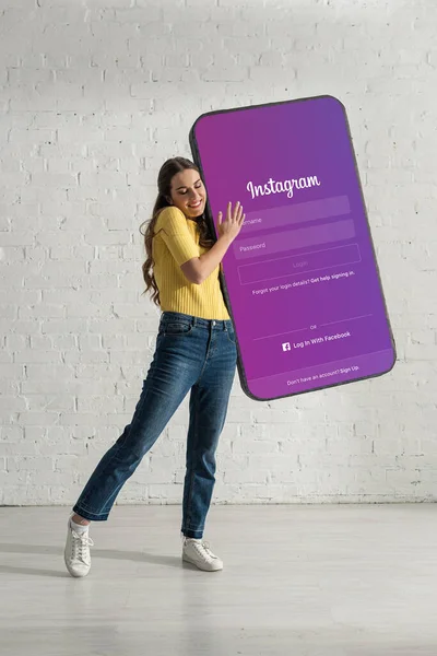 KYIV, UKRAINE - FEBRUARY 21, 2020: Attractive woman smiling while holding huge model of smartphone with instagram app near white brick wall — Stock Photo