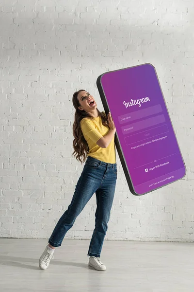 KYIV, UKRAINE - FEBRUARY 21, 2020: Cheerful girl laughing while holding big model of smartphone with instagram app — Stock Photo