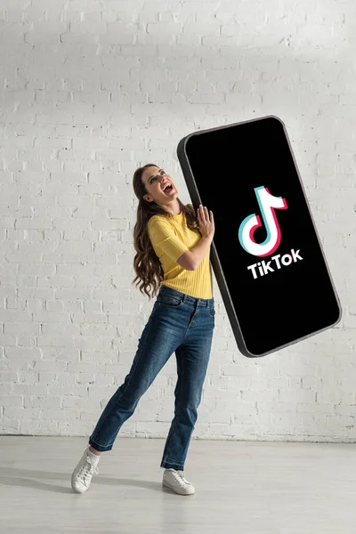 KYIV, UKRAINE - FEBRUARY 21, 2020: Attractive woman laughing while holding huge model of smartphone with TikTok app near white brick wall — Stock Photo