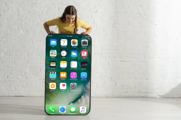 KYIV, UKRAINE - FEBRUARY 21, 2020: Girl looking at big model of smartphone with iphone screen near white brick wall — Stock Photo