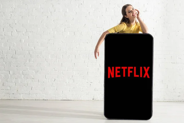 KIEW, UKRAINE - 21. FEBRUAR 2020: Schöne Frau mit Hand in der Nähe der Wange, die in die Kamera lächelt, in der Nähe eines großen Smartphone-Modells mit Netflix-App — Stockfoto