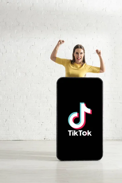 KYIV, UKRAINE - FEBRUARY 21, 2020: Cheerful girl showing yeah gesture while standing near model of smartphone with TikTok app — Stock Photo