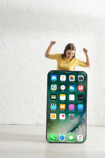 KYIV, UKRAINE - FEBRUARY 21, 2020: Positive girl looking at big model of smartphone with iphone screen while showing yeah gesture — Stock Photo