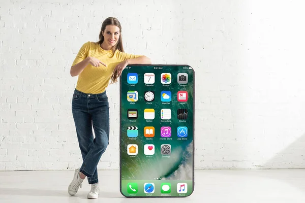 KYIV, UCRANIA - 21 de febrero de 2020: Muchacha sonriente que señala con el dedo al modelo de teléfono inteligente con pantalla de iphone - foto de stock