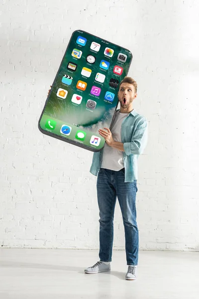 KYIV, UCRANIA - 21 de febrero de 2020: Hombre sorprendido sosteniendo gran modelo de teléfono inteligente con pantalla de iphone en casa - foto de stock