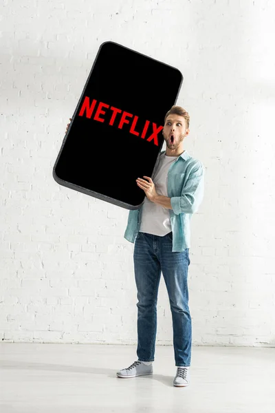 KYIV, UCRANIA - 21 de febrero de 2020: Emocionado hombre sosteniendo el modelo de teléfono inteligente con aplicación netflix cerca de la pared de ladrillo blanco — Stock Photo