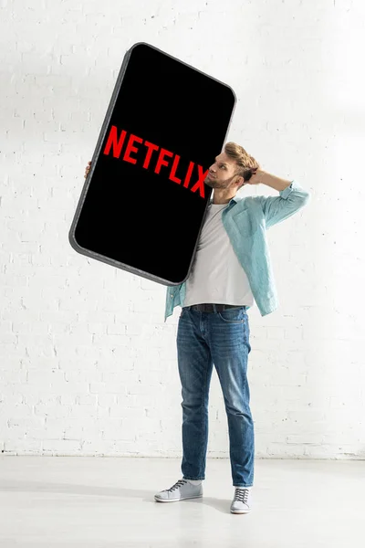 KIEW, UKRAINE - 21. FEBRUAR 2020: Nachdenklicher Mann mit händennahem Kopf, der ein großes Smartphone-Modell mit Netflix-App hält — Stockfoto
