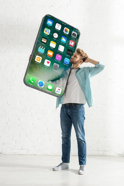 KYIV, UCRANIA - 21 de febrero de 2020: Hombre pensativo mirando gran modelo de teléfono inteligente con pantalla de iphone en casa - foto de stock