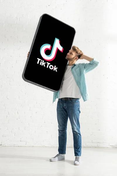 KYIV, UKRAINE - FEVEREIRO 21, 2020: Homem pensativo segurando modelo de smartphone com aplicativo TikTok perto da parede de tijolo branco — Fotografia de Stock