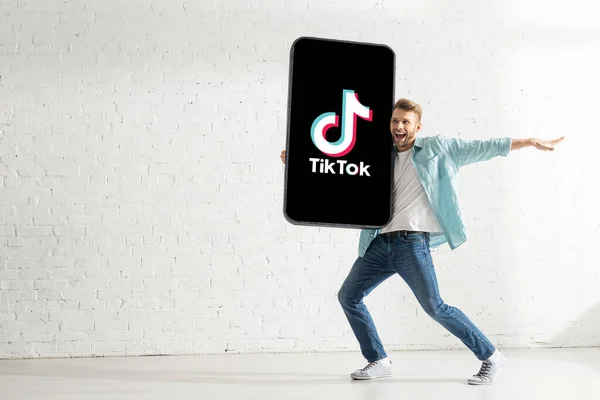 KYIV, UKRAINE - 21 février 2020 : Un bel homme souriant tout en tenant un grand modèle de smartphone avec l'application TikTok à la maison — Photo de stock