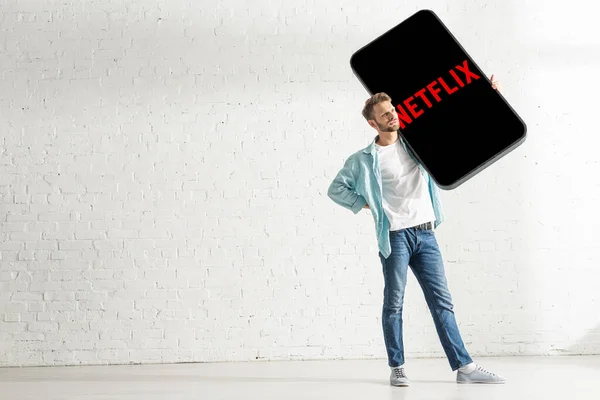 KYIV, UKRAINE - FEBRUARY 21, 2020: Handsome man with hand on hip holding big model of smartphone with netflix app at home — Stock Photo