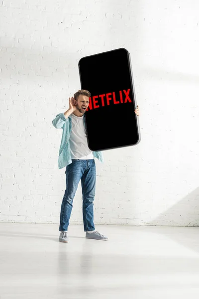 KYIV, UKRAINE - FEBRUARY 21, 2020: Handsome man with hand near ear holding smartphone model with netflix app near white brick wall — стокове фото