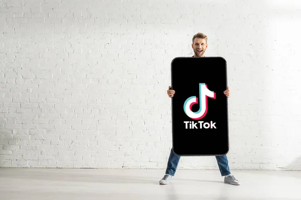 KYIV, UCRANIA - 21 de febrero de 2020: Hombre guapo sosteniendo un gran modelo de teléfono inteligente con la aplicación TikTok y sonriendo a la cámara - foto de stock