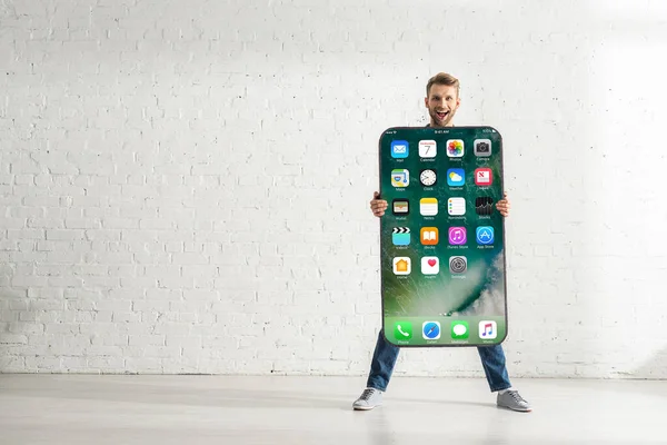 KYIV, UKRAINE - FEBRUARY 21, 2020: Cheerful man holding smartphone model with iphone screen near white brick wall — Stock Photo