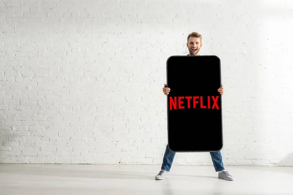 KYIV, UKRAINE - FEBRUARY 21, 2020: Handsome man smiling at camera while holding big model of smartphone with netflix app at home — Stock Photo