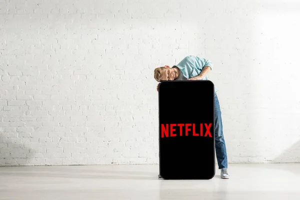 KYIV, UKRAINE - 21 FÉVRIER 2020 : Homme souriant debout près de grand modèle de smartphone avec application netflix à la maison — Photo de stock