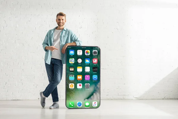 KYIV, UKRAINE - FEBRUARY 21, 2020: Handsome man smiling at camera and pointing with finger at big model of smartphone with iphone screen — Stock Photo