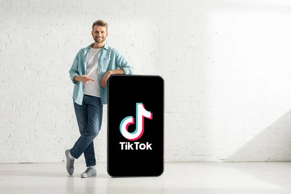 KYIV, UKRAINE - FEBRUARY 21, 2020: Handsome man pointing with finger at model of smartphone with TikTok app and smiling at camera — Stock Photo