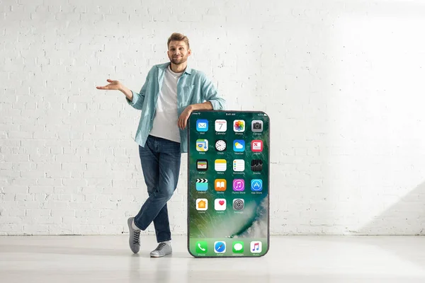 KYIV, UCRANIA - 21 de febrero de 2020: Hombre guapo sonriendo mientras muestra un gesto de dubio cerca de un gran modelo de teléfono inteligente con pantalla de iphone - foto de stock